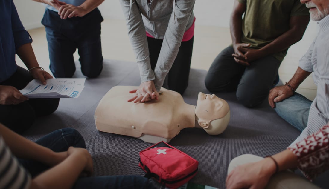 Adults taking part in a first aid training session, practising CPR on a manikin with a first aid kit nearby.