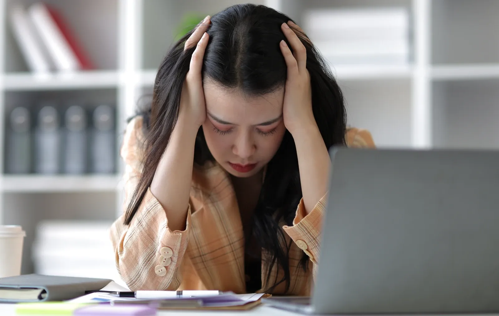 Office worker holding her head while working at a laptop, illustrating workplace stress and workload pressure.