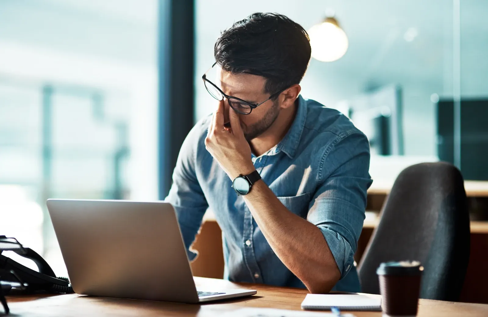 Office worker sitting at a desk with one hand over his face, looking tired and stressed while working on a laptop