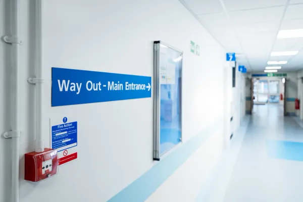 NHS hospital corridor with blue “Way Out – Main Entrance” sign and fire alarm on wall.
